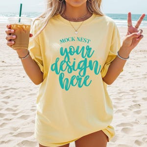 May include: A light yellow t-shirt with the text "MOCK NEST your design here" in teal script. The person is holding a clear plastic cup with a straw and iced coffee. The background is a sandy beach.