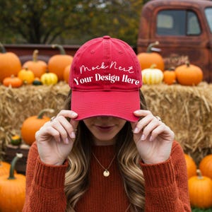 May include: A red baseball cap with the text "Mock Nest Your Design Here" in white script. The cap is held up in front of a person's face. Pumpkins and hay bales are in the background, suggesting an autumn theme.