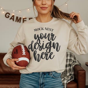 Könnte beinhalten: Ein beigefarbenes Sweatshirt mit dem Text "MOCK NEST your design here" in schwarzer Schrift. Ein brauner und weißer Football wird in den Händen der Person gehalten. Das Wort "GAME" ist im Hintergrund zu sehen.