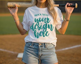 May include: White t-shirt with the text "MOCK NEST your design here" in teal script. The person is wearing denim shorts and holding a baseball bat. The background is a baseball field.