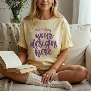以下が含まれることがあります： 紫色のスクリプトで「MOCK NEST your design here」というテキストがプリントされた、袖をまくった薄い黄色のTシャツ。モデルはソファに座り、本を持ち、ショートパンツとクリーム色の靴下を履いています。