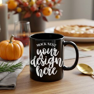 Op de afbeelding: Een zwarte keramische mok met een gebogen handvat. De mok heeft de tekst "MOCK NEST" boven de woorden "your design here" in een wit script. Een oranje pompoen en een taart staan op de achtergrond.