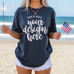 May include: Dark gray t-shirt with the text "MOCK NEST your design here" in white script. The person is holding a sparkler and a small American flag. The person is wearing white shorts with a fringed hem. The background is a beach.