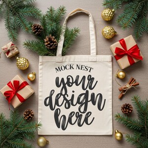 May include: A cream-colored tote bag with the text "MOCK NEST your design here" in black script. The bag is surrounded by Christmas decorations, including wrapped gifts with red bows, pine needles, pine cones, and gold ornaments.
