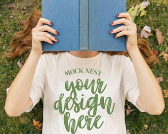 May include: A person lying on grass, holding a blue book in front of their face. They are wearing a cream-colored t-shirt with the text "MOCK NEST your design here" in green script. The person is also wearing blue jeans.