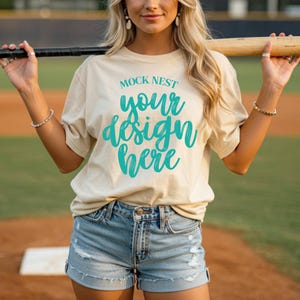 May include: A beige t-shirt with the text "MOCK NEST your design here" in teal script. The model is wearing denim shorts and holding a baseball bat. The background is a baseball field.