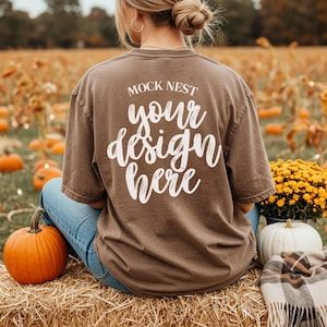 以下が含まれることがあります： 「MOCK NEST your design here」の白文字がプリントされた茶色のTシャツ。Tシャツは、カボチャと秋の葉を背景に、干し草の俵に座っている人が着ています。