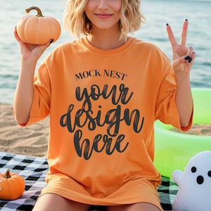 May include: Orange t-shirt with the text "MOCK NEST your design here". The image includes a small orange pumpkin and a white ghost-shaped plush toy. The person is holding a small orange pumpkin. The background is a beach setting.