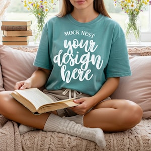 May include: Teal t-shirt with the text "MOCK NEST your design here" in white script. The person is wearing beige shorts and white socks. A book is open in their lap. Flowers and books are in the background.