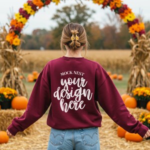 以下が含まれることがあります： 白文字で「MOCK NEST your design here」と書かれたマルーンのスウェットシャツ。秋をテーマにしたアーチの前で、カボチャと干し草の俵があります。髪は金のヘアクリップでスタイリングされています。
