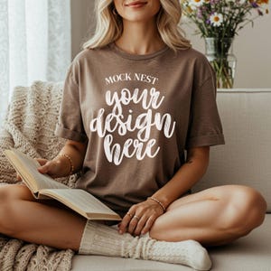 May include: A brown t-shirt with rolled-up sleeves featuring the text "MOCK NEST your design here" in white script. The person is sitting on a couch and reading a book. The person is wearing white socks.