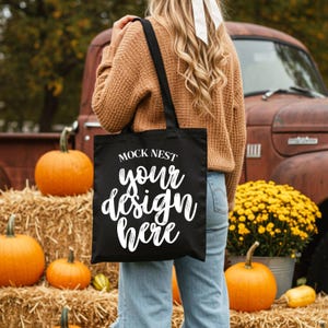 May include: A black tote bag with the text "MOCK NEST your design here" in white script. The bag is held in front of a fall scene with pumpkins, hay bales, and a vintage truck. The bag has black handles.