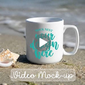 May include: A white ceramic mug with a large handle, featuring the text "MOCK NEST your design here" in teal script. A play button graphic is in the center. The words "Video Mock-up" are at the bottom. A seashell rests beside the mug.