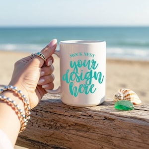 May include: A white ceramic mug with the text "MOCK NEST your design here" in teal script. The mug is held by a hand adorned with a silver bracelet and rings. A seashell and a piece of green sea glass rest on a wooden surface.
