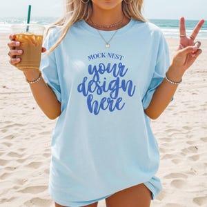 Pode incluir: Camiseta azul clara com o texto "MOCK NEST your design here" em escrita azul. A camiseta é usada em uma praia com o oceano ao fundo. A pessoa está segurando um copo transparente com um canudo e café gelado.
