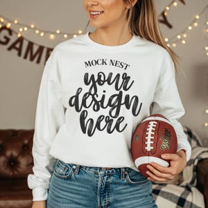 May include: A white sweatshirt with the text "MOCK NEST your design here" in black script. The person is holding a brown and white football. The person is wearing blue jeans.