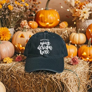 May include: A black baseball cap with the text "MOCK NEST your design here" in white script. The cap is displayed on a hay bale, surrounded by carved pumpkins, flowers, and string lights, creating a festive autumn scene.