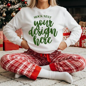 May include: White long-sleeve sweatshirt with the text "MOCK NEST your design here" in green. The person is wearing red and white plaid pajama pants and white socks. The scene includes a Christmas tree and wrapped gifts.