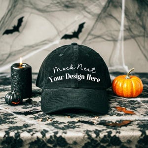 May include: A black corduroy baseball cap with the text "Mock Nest Your Design Here". The image features Halloween decorations, including a lit candle, pumpkins, and bat silhouettes.