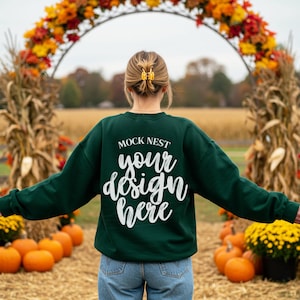 Puede incluir: Una sudadera verde oscuro con el texto "MOCK NEST your design here" en escritura blanca. La sudadera se usa al aire libre con calabazas y follaje de otoño en el fondo.