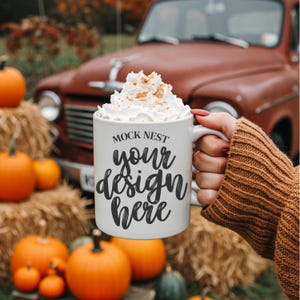 Pode incluir: Uma caneca de café branca com o texto "MOCK NEST your design here" é preenchida com chantilly e canela. A caneca é segurada em frente a uma cena de outono com abóboras, fardos de feno e uma caminhonete vermelha vintage.