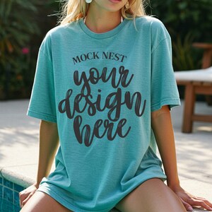 Op de afbeelding: Een turquoise t-shirt met de tekst "MOCK NEST your design here" in zwarte letters. Het shirt heeft een relaxte pasvorm en lijkt van zacht materiaal te zijn. De persoon zit bij een zwembad.
