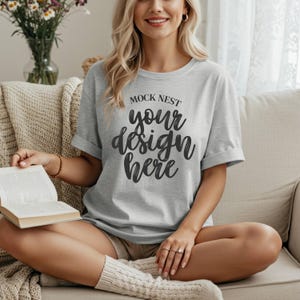 May include: A light gray t-shirt with rolled-up sleeves featuring the text "MOCK NEST your design here" in a black script font. The model is sitting on a beige couch, wearing beige shorts and white socks.