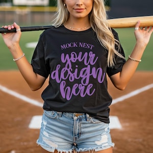 May include: Black t-shirt with the text "MOCK NEST your design here" in purple script. The model is wearing denim shorts and holding a baseball bat. The background is a baseball field.