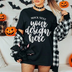 Pode incluir: Camiseta preta com o texto "MOCK NEST your design here" em escrita branca. A modelo está usando um colar gargantilha preto e um cardigan xadrez preto e branco. Um brinquedo de pelúcia de morcego é segurado na mão da modelo. Tema de Halloween.