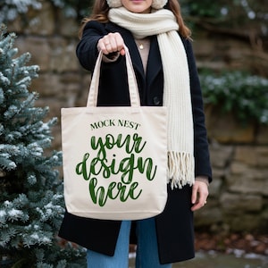 May include: A cream-colored tote bag with the text "MOCK NEST your design here" in green script. The bag is held by a person wearing a black coat, scarf, and earmuffs. The background includes a stone wall and a snowy evergreen tree.