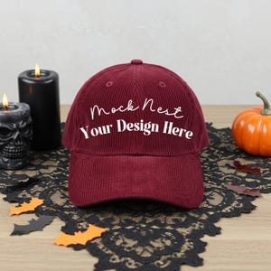 May include: A burgundy corduroy baseball cap with the text "Mock Nest Your Design Here" in white. The cap is displayed on a black lace runner with Halloween decorations, including a pumpkin and candles.