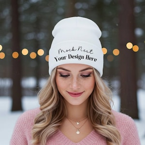 May include: A white beanie with the text "Mock Nest Your Design Here" in black script. The beanie is worn by a person with blonde hair and a pink sweater. The background is blurred with bokeh lights.