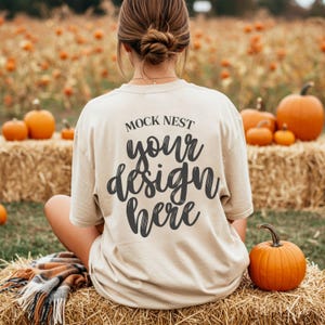 May include: A beige t-shirt with the text "MOCK NEST your design here" in black script. The shirt is worn outdoors with pumpkins and hay bales in the background. The shirt is a mock-up for a design.