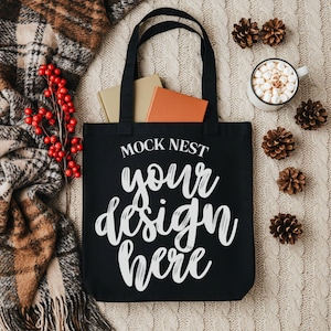 May include: Black tote bag with the text "MOCK NEST your design here". The bag contains two books. The image also includes a mug of hot chocolate with marshmallows, pine cones, a plaid blanket, and red berries.