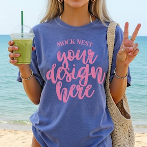May include: A blue t-shirt with the text "MOCK NEST your design here" in pink. The person is holding a green drink in a clear plastic cup with a green straw. A beige woven bag is over the shoulder.