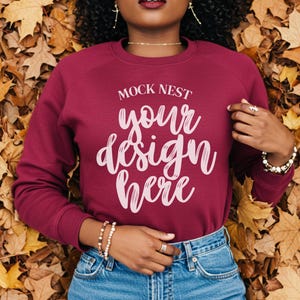 以下が含まれることがあります： ライトピンクのスクリプトで「MOCK NEST your design here」というテキストが入ったマルーンのスウェットシャツ。モデルはブルージーンズとジュエリーを着用しています。背景は秋の葉です。