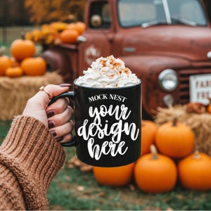Op de afbeelding: Een zwarte koffiemok met de tekst "MOCK NEST your design here" in witte letters. De mok is gevuld met slagroom en kaneel. De achtergrond omvat pompoenen, hooibalen en een vintage rode vrachtwagen.