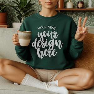 Puede incluir: Una sudadera verde oscuro con el texto "MOCK NEST your design here" en escritura blanca. La persona sostiene una taza beige y marrón. La persona lleva pantalones cortos beige y calcetines blancos.