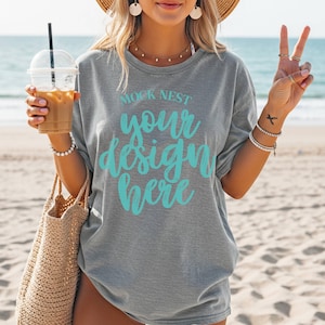May include: Gray t-shirt with the text "MOCK NEST your design here" in turquoise. The model is holding a clear plastic cup with a straw and a woven tote bag. The background is a beach with the ocean in the distance.