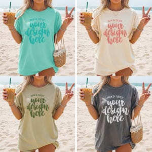 May include: Four t-shirts in different colors: teal, cream, olive green, and charcoal gray. Each shirt has the text "MOCK NEST your design here" in a script font. The shirts are worn on a beach setting.