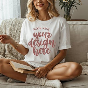 Könnte beinhalten: Weißes T-Shirt mit hochgekrempelten Ärmeln und dem Text "MOCK NEST your design here" in einer Schreibschrift. Das T-Shirt wird von einer Person getragen, die auf einem Sofa sitzt und ein Buch hält. Das Thema ist Freizeitkleidung.