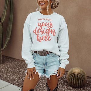 May include: A light blue sweatshirt with the text "MOCK NEST your design here" in coral lettering. The model is wearing denim shorts, a brown belt with a decorative buckle, and cowboy boots. A cactus is in the background.