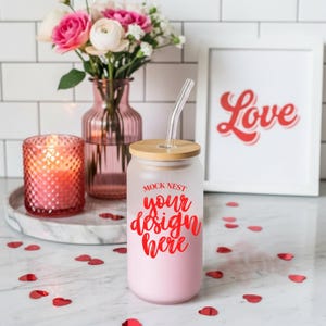 May include: A pink glass tumbler with a bamboo lid and a clear straw. The tumbler has the text "Mock Nest your design here" in red. A vase of pink flowers, a candle, and a framed print that says "Love" are in the background.