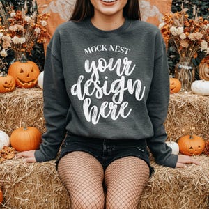May include: Dark gray crewneck sweatshirt with the text "MOCK NEST your design here" in white script. The sweatshirt is part of a fall-themed display with pumpkins and hay bales.