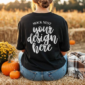 May include: Black t-shirt with the text "MOCK NEST your design here" in white script. The person is sitting on a hay bale with pumpkins and flowers nearby. The background is a blurred field.