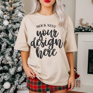 Könnte beinhalten: Beiges T-Shirt mit dem Text "MOCK NEST your design here" in schwarzer Schrift. Das Model trägt einen rot-grünen karierten Rock. Ein Weihnachtsbaum und ein Kamin befinden sich im Hintergrund.