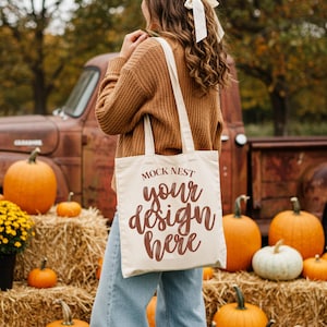 May include: Cream-colored tote bag with the text "MOCK NEST your design here" in brown script. The bag is carried in front of a rustic truck and pumpkins. The scene evokes a fall harvest theme.