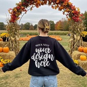 Puede incluir: Una sudadera negra con el texto "MOCK NEST your design here" en escritura blanca. La persona está en un campo de calabazas con balas de heno y un arco decorado con follaje de otoño y calabazas.
