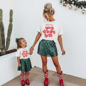May include: Two people in matching outfits. Both wear off-white t-shirts with the text "MOCK NEST your design here" in red. They also wear green sequin skirts and red cowboy boots with white detailing. A child holds the adult's hand. The outfits are festive.