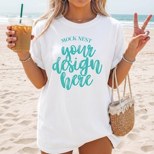Può includere: T-shirt bianca con la scritta "MOCK NEST your design here" in corsivo turchese. La modella è su una spiaggia, con in mano una bevanda e una piccola borsa di paglia. La modella fa anche il segno della pace.